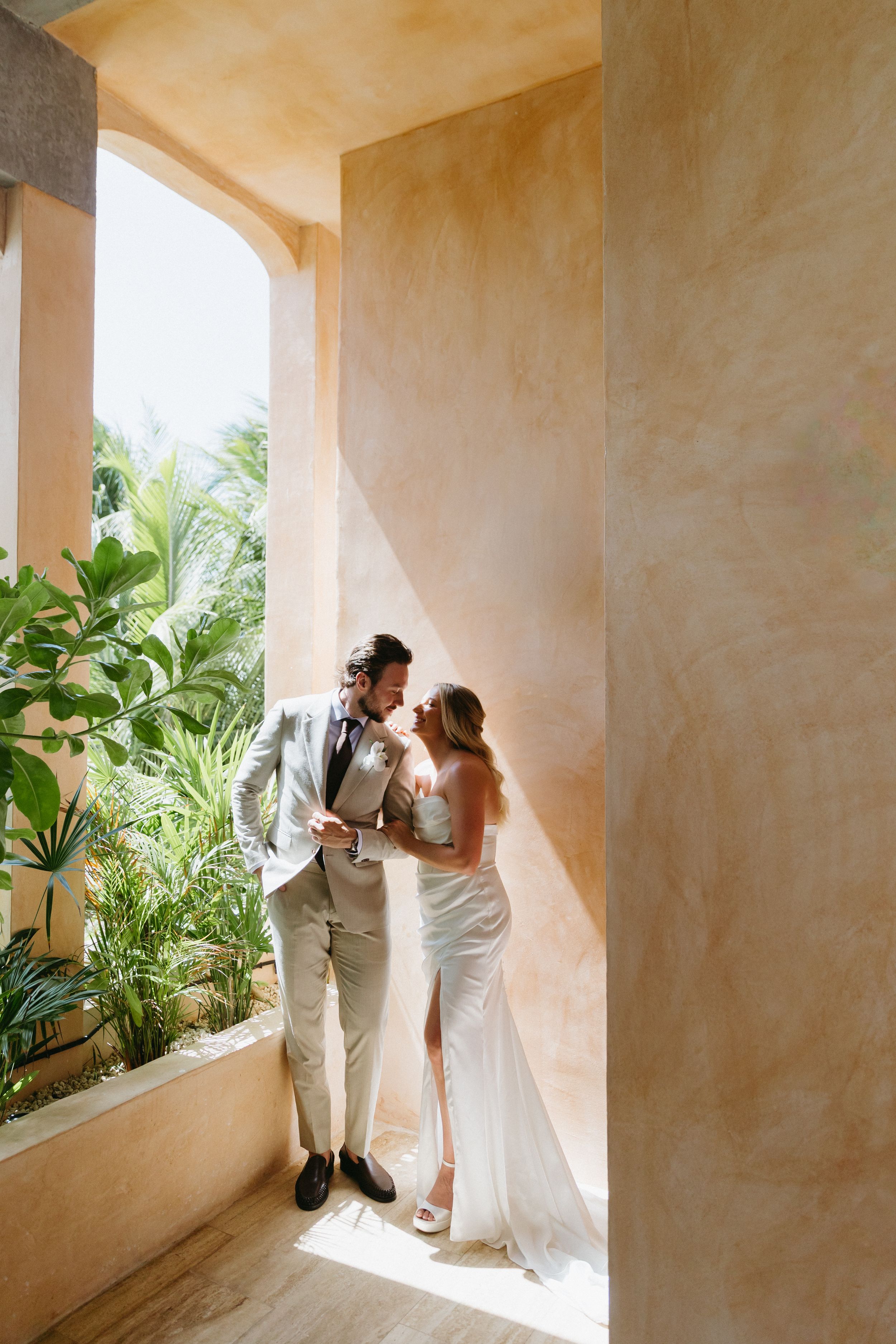 Bride and groom share a romantic moment in a sunlit archway surrounded by tropical greenery at a destination wedding.