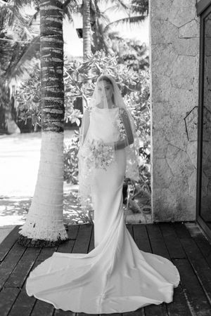 Elegant bride in sleek white gown with long train and veil posing beside a tropical palm tree.