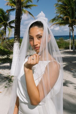 Beautiful bride in white gown and veil poses near palm trees on sandy beach, showcasing elegant engagement ring.
