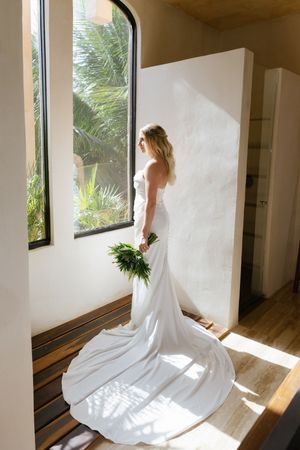 Elegant bride in a strapless white gown with a long train holds a green bouquet by a sunlit arched window.