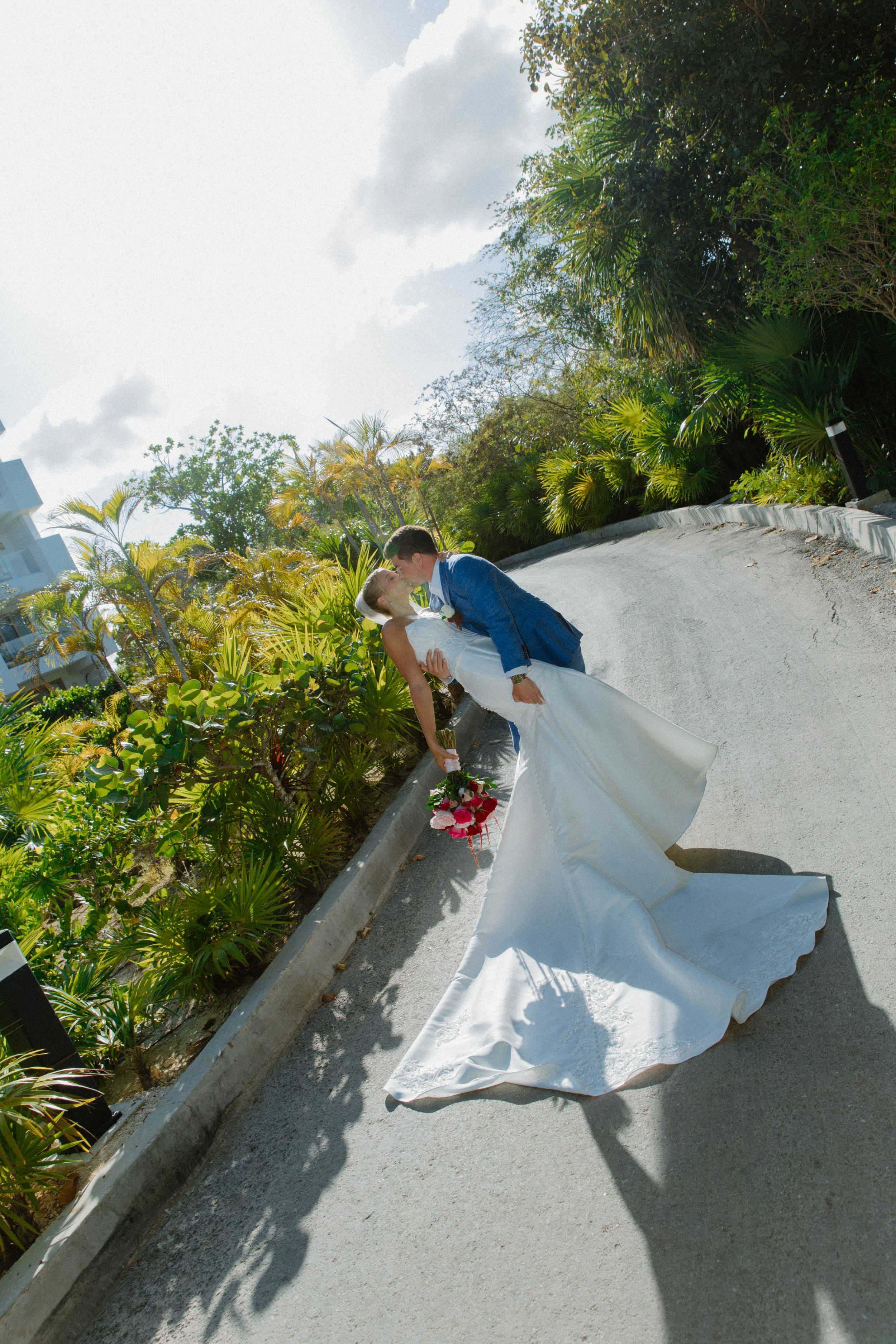 Bride and groom sharing a romantic dip kiss on a curved pathway surrounded by lush tropical greenery.