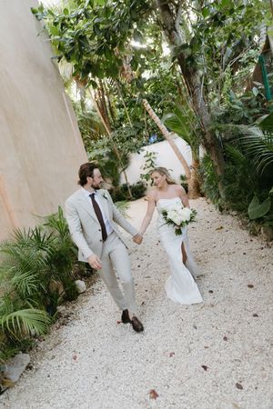 Bride and groom walk hand-in-hand on a gravel path surrounded by lush tropical greenery during their destination wedding.