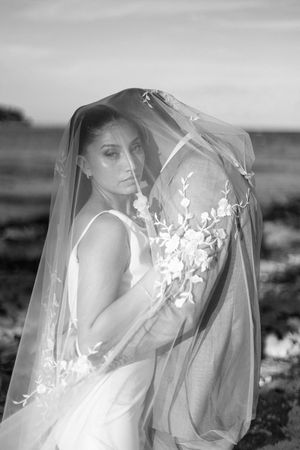 Bride draped in floral embroidered veil poses outdoors in black and white wedding portrait.