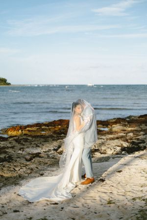 Bride and groom share romantic moment under veil on rocky beach with ocean backdrop.
