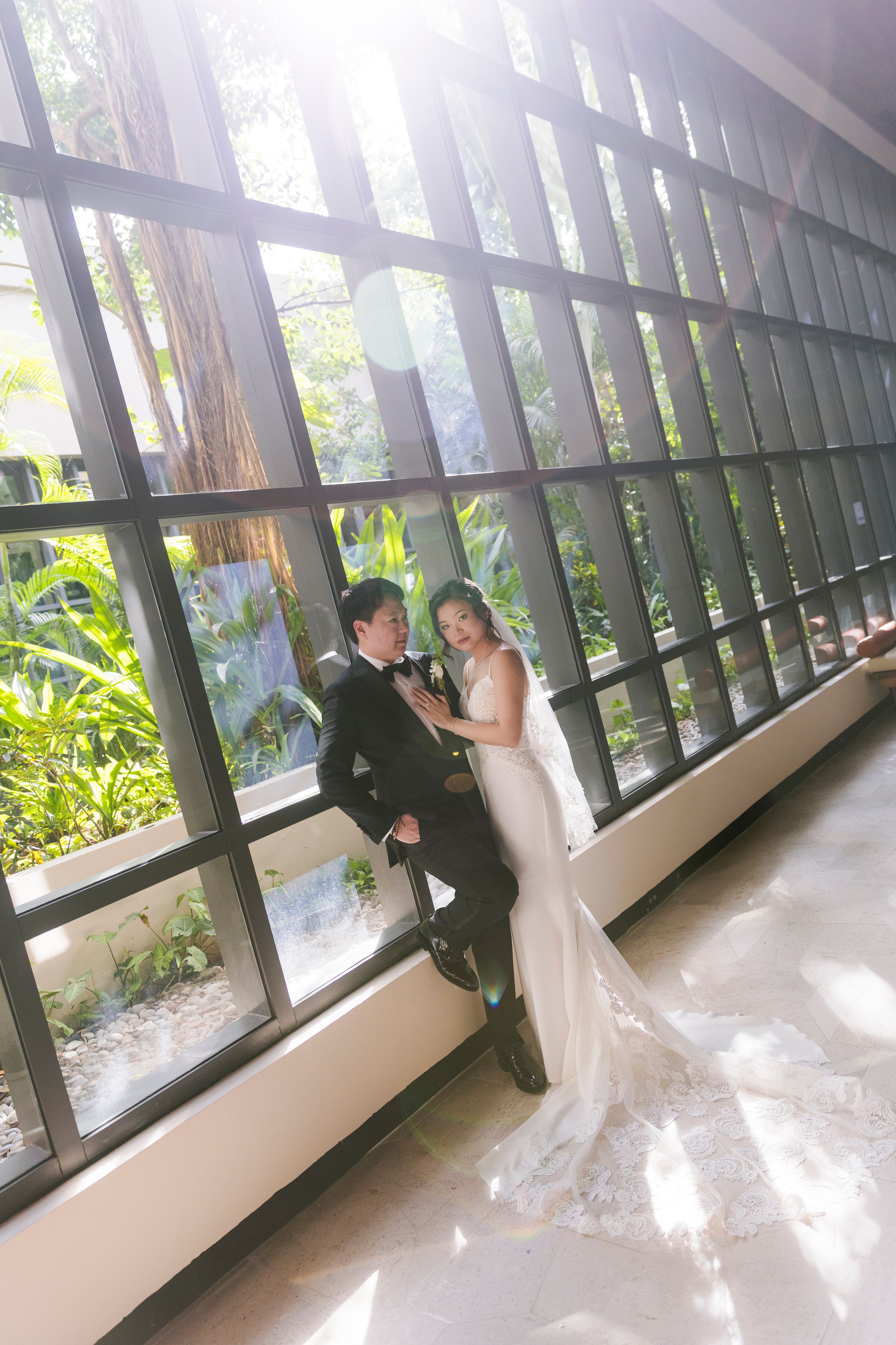 Bride and groom sharing an intimate moment in a sunlit corridor with floor-to-ceiling windows.