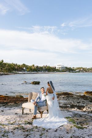 Bride and groom pose on rocky shoreline with tropical resort and blue sky in background.