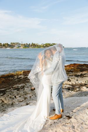 Bride and groom share a romantic moment under a floral veil on a rocky beach with ocean views.