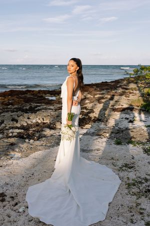 Bride in white gown with train holds bouquet on rocky beach shore with ocean backdrop.