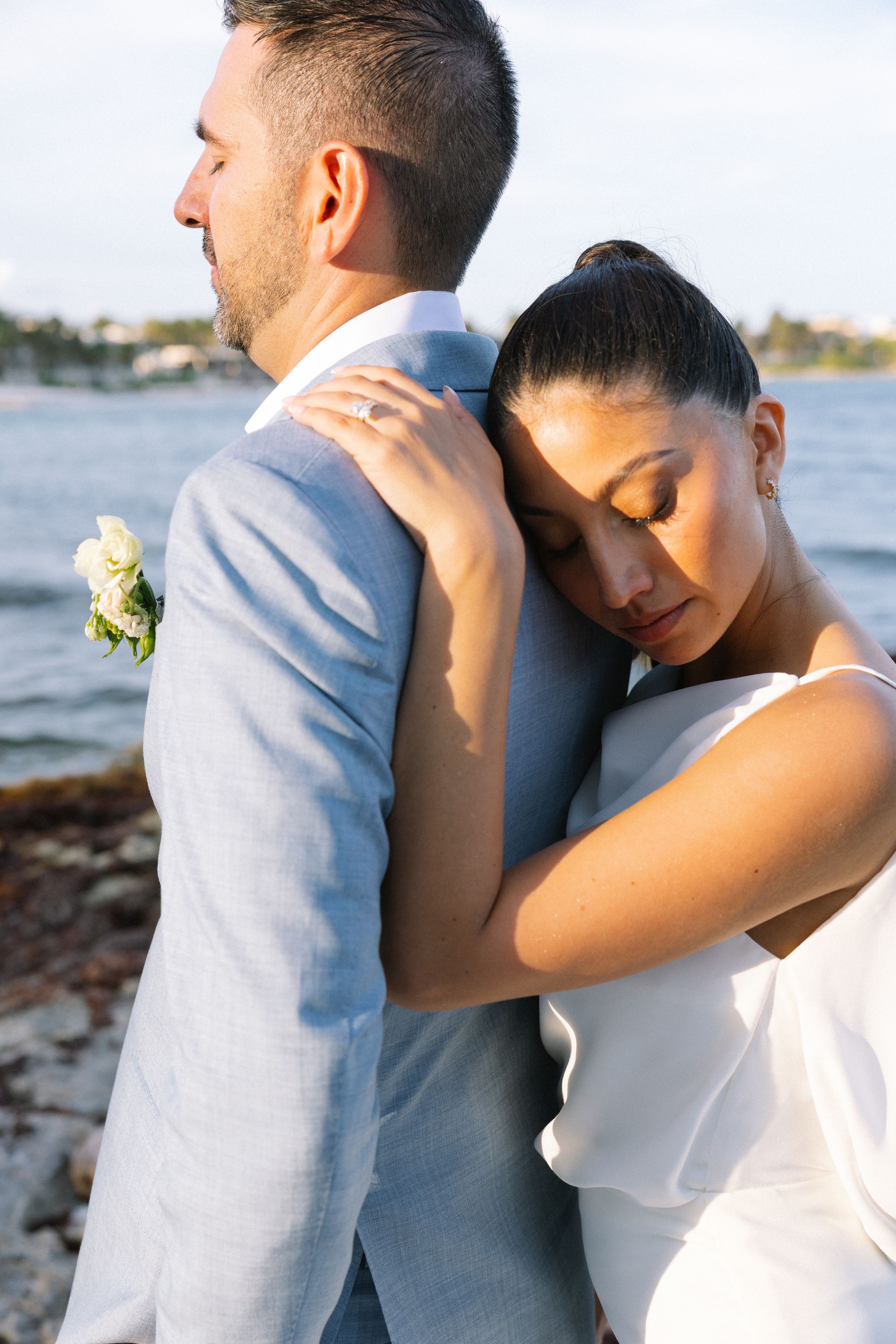 Bride in white dress rests head on groom's shoulder in blue suit near waterfront at golden hour.
