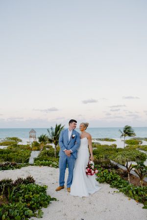 Bride and groom pose on sandy beach path with ocean backdrop at sunset destination wedding.