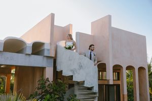 Bride and groom stand atop curved stairs of a modern stucco building at a tropical destination wedding venue.