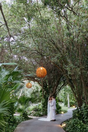 Bride and groom embrace on a lush garden path under glowing wicker lanterns and canopy trees.