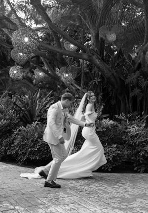 Black and white photo of a bride and groom dancing joyfully outdoors amid lush tropical greenery and decorative globe lights.