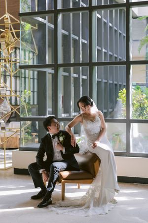 Groom seated in leather chair gazing at bride in lace wedding gown in a modern sunlit interior.