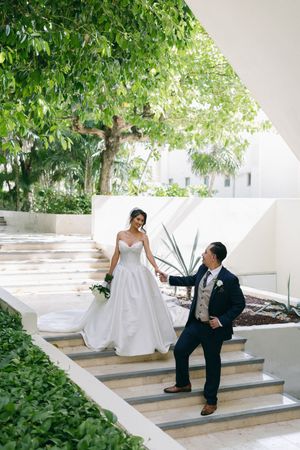 Bride in white gown and groom in navy suit pose on outdoor steps surrounded by lush greenery at a resort wedding.