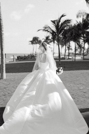 Bride in elegant white ball gown and veil holding bouquet, standing near palm trees on a beach in black and white photo.