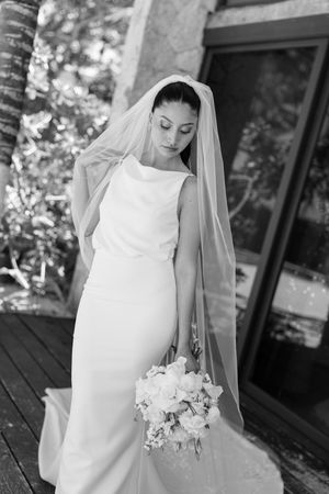 Elegant bride in white gown and veil holding floral bouquet, black and white wedding portrait outdoors.