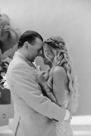 Black and white photo of newlyweds sharing intimate forehead touch moment during outdoor tropical wedding ceremony.