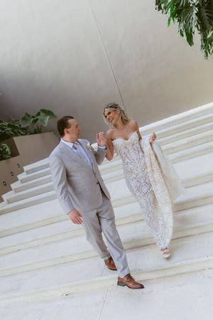 Bride in lace gown holds train while sharing a smile with groom in gray suit on elegant white marble staircase.