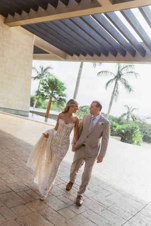 Newlyweds walk hand-in-hand in lace wedding gown and gray suit under modern pergola with tropical palm trees.