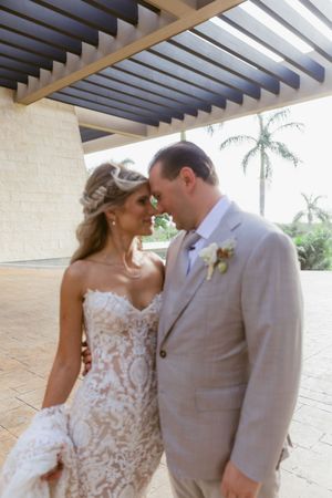 Bride in intricate lace gown and groom in gray suit share romantic nose-to-nose moment under tropical resort pergola.