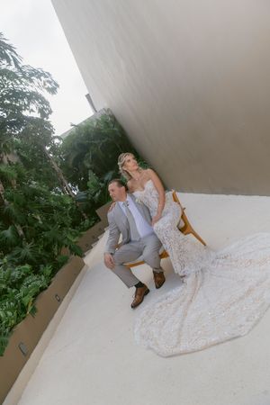 Bride in flowing lace gown and groom in gray suit sit together on balcony surrounded by lush tropical greenery.