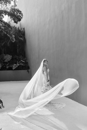 Bride in elegant wedding gown with flowing cathedral veil billowing in the wind against a neutral wall.