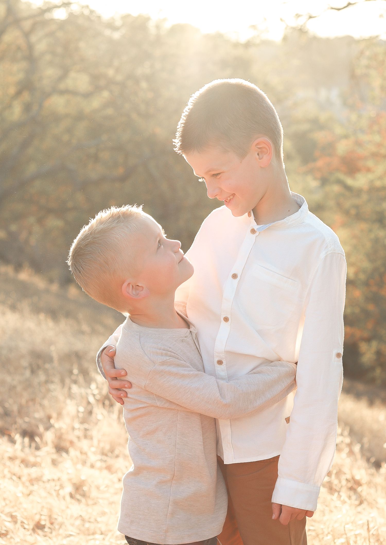 Two young boys hugging and smiling at each other in a sunny golden field during a warm outdoor photo session.