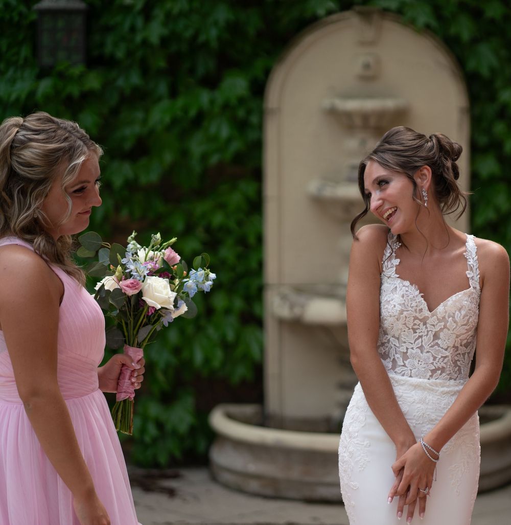 Bride in lace gown smiling at bridesmaid in pink dress holding bouquet, with stone fountain backdrop.