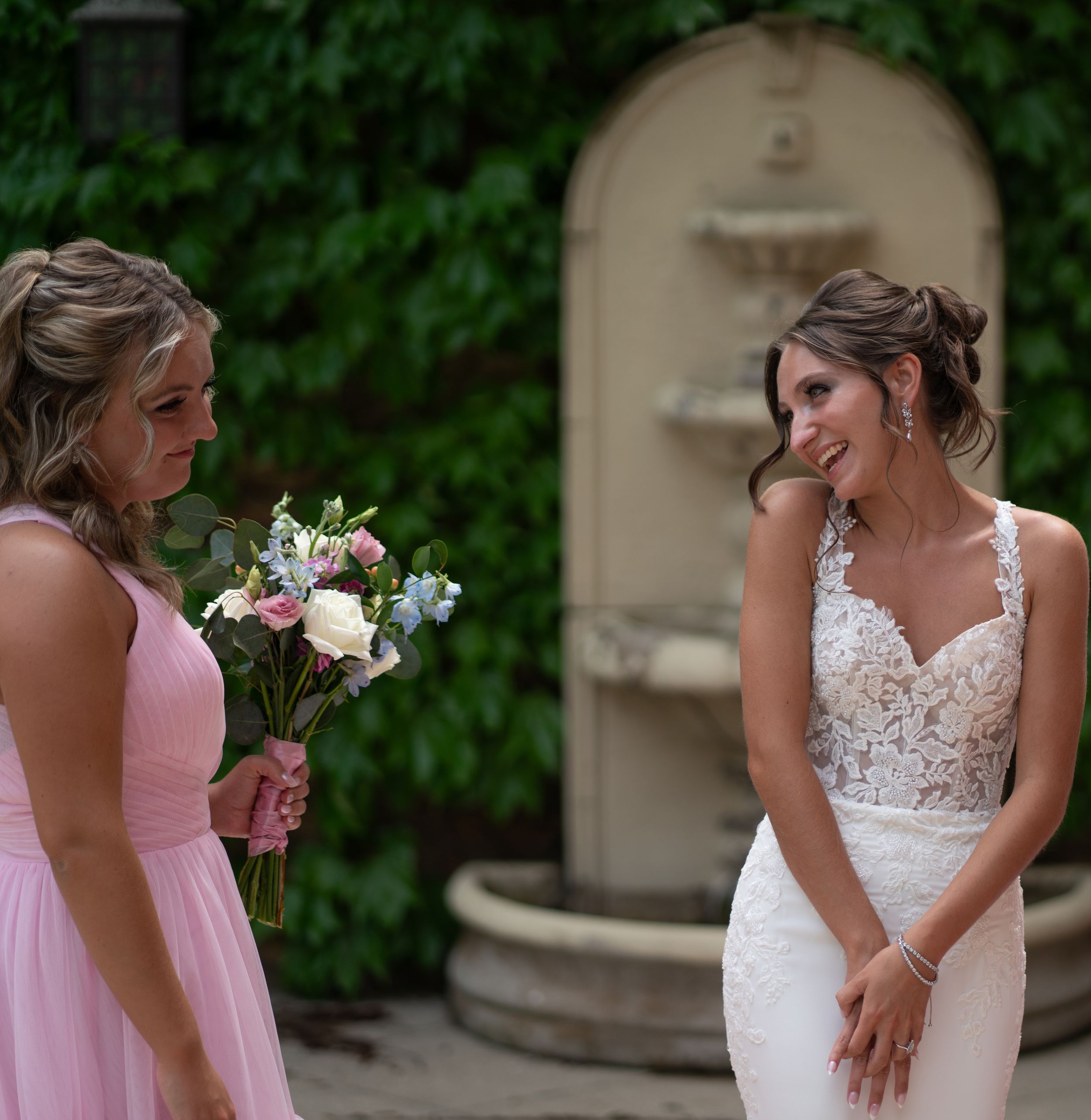 Bride in lace gown smiling at bridesmaid in pink dress holding bouquet, with stone fountain backdrop.