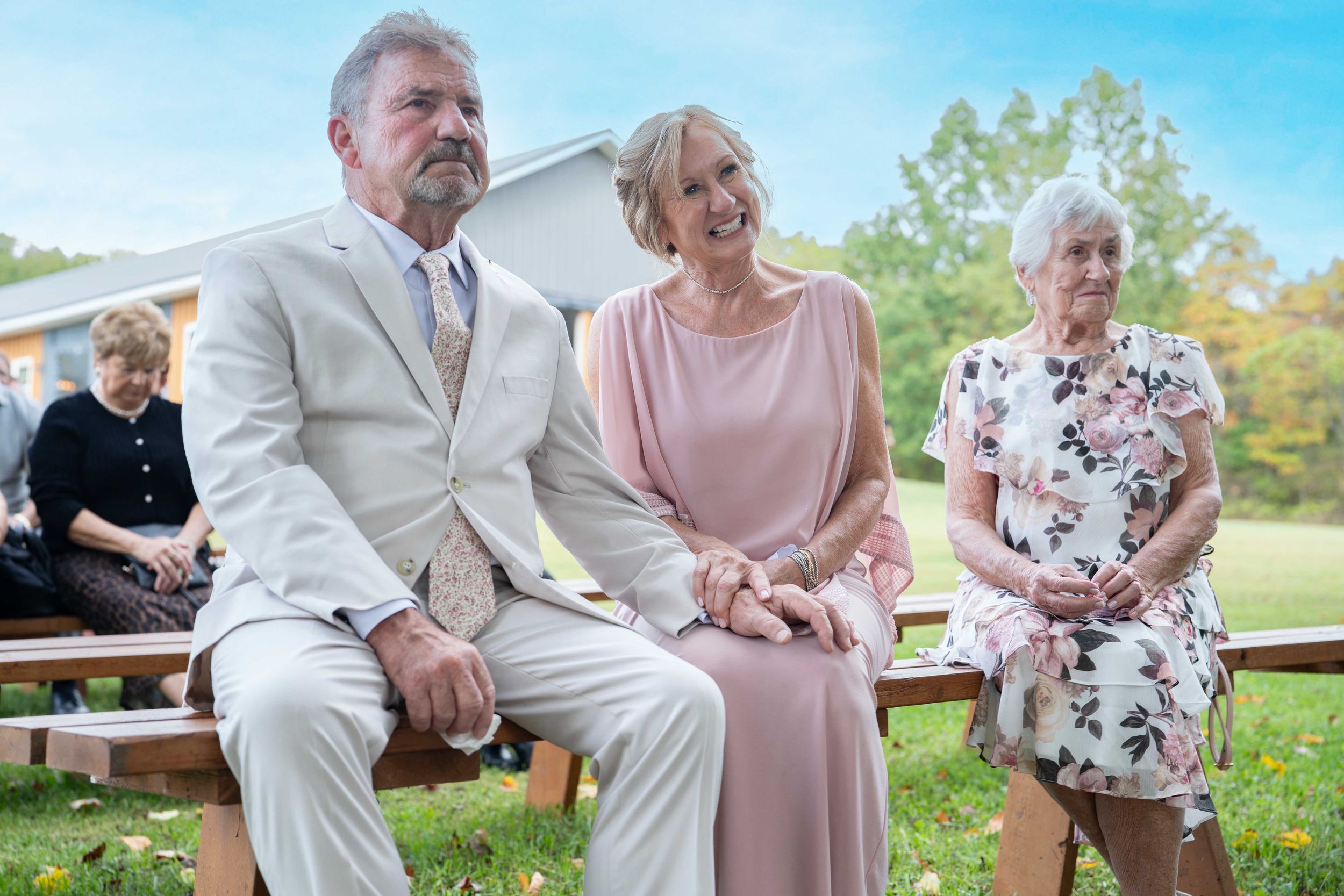 Elderly couple in formal attire sitting on wooden bench at outdoor wedding ceremony on sunny day.