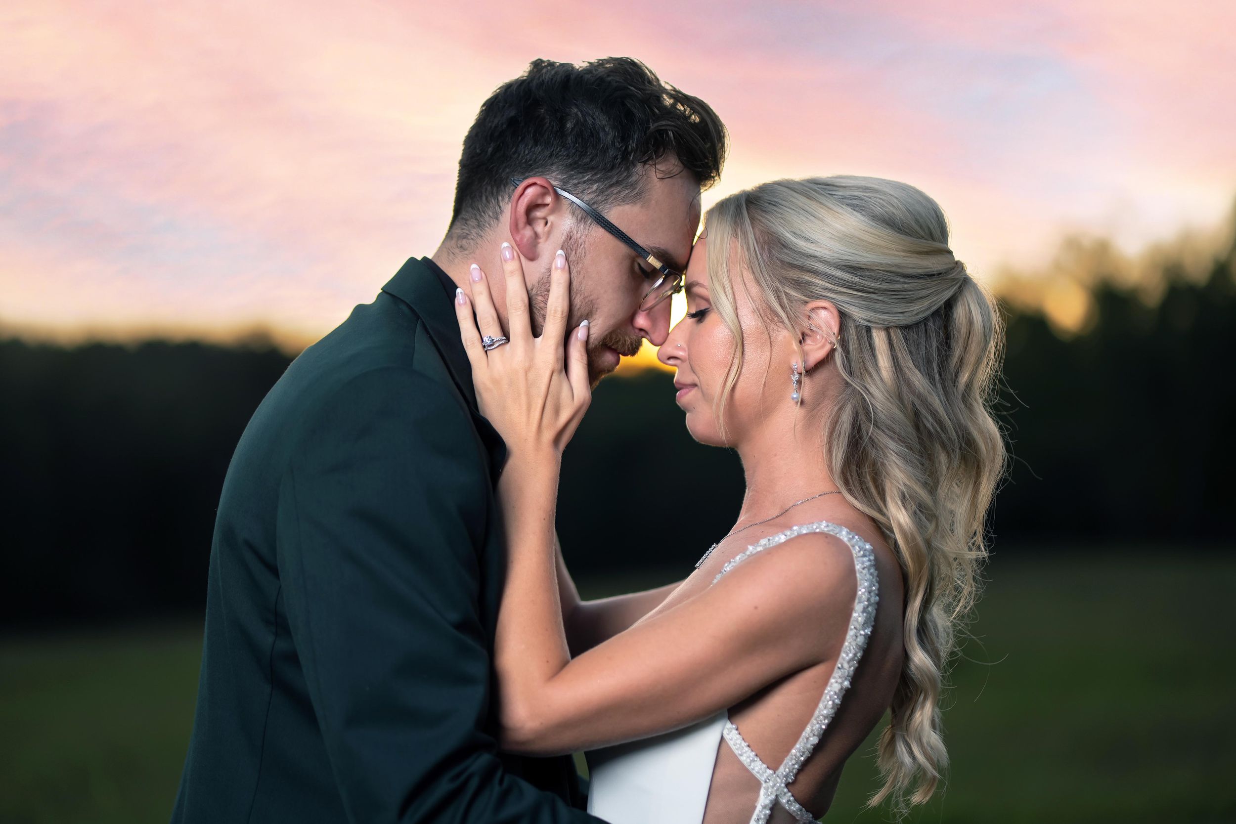 Romantic couple sharing an intimate moment at sunset with a colorful pink sky backdrop at their outdoor wedding.