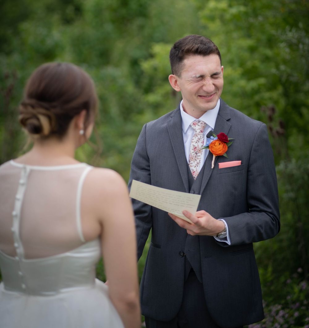 Groom smiling while reading wedding vows to bride outdoors, wearing gray suit with floral boutonniere.