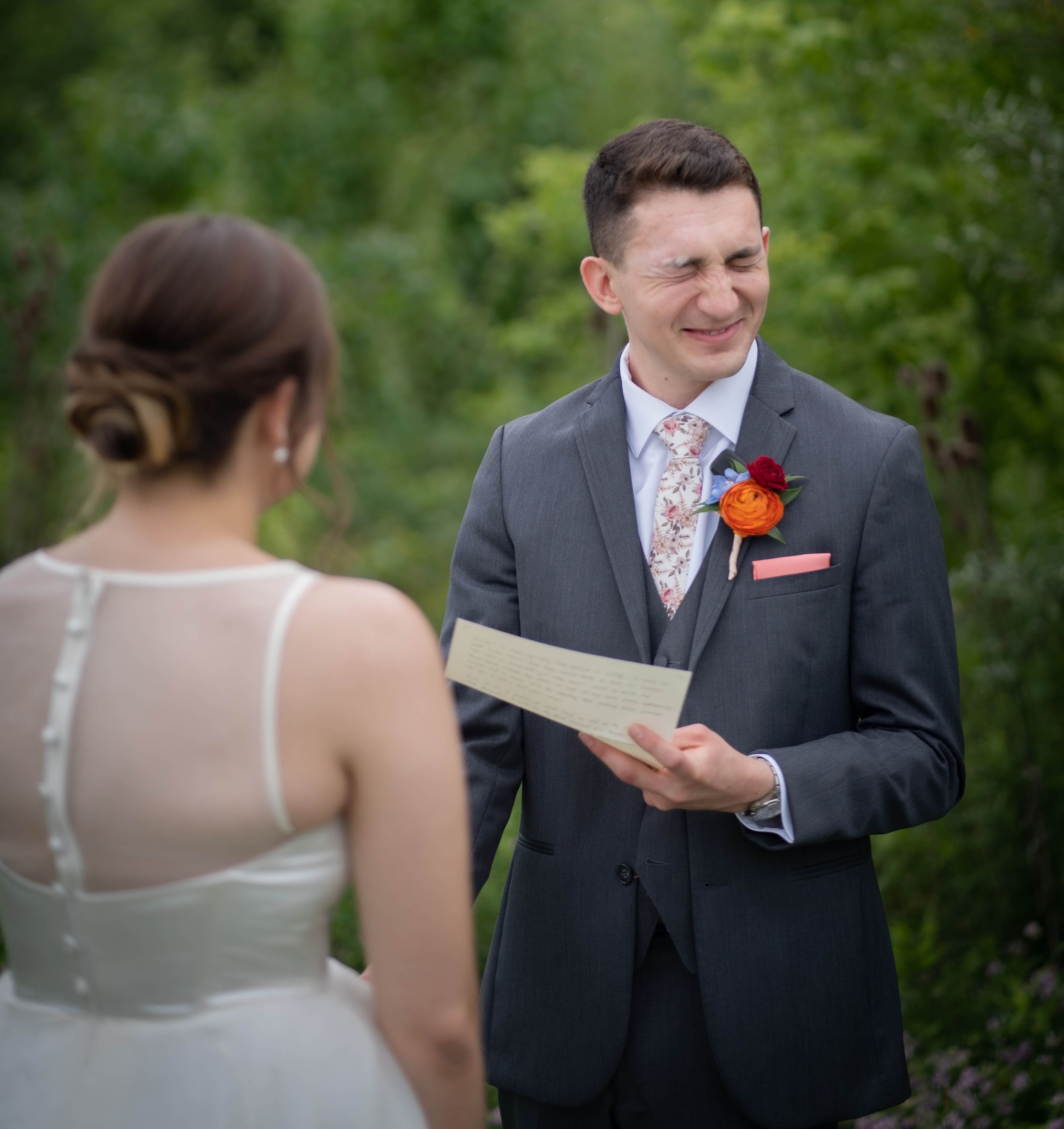 Groom smiling while reading wedding vows to bride outdoors, wearing gray suit with floral boutonniere.