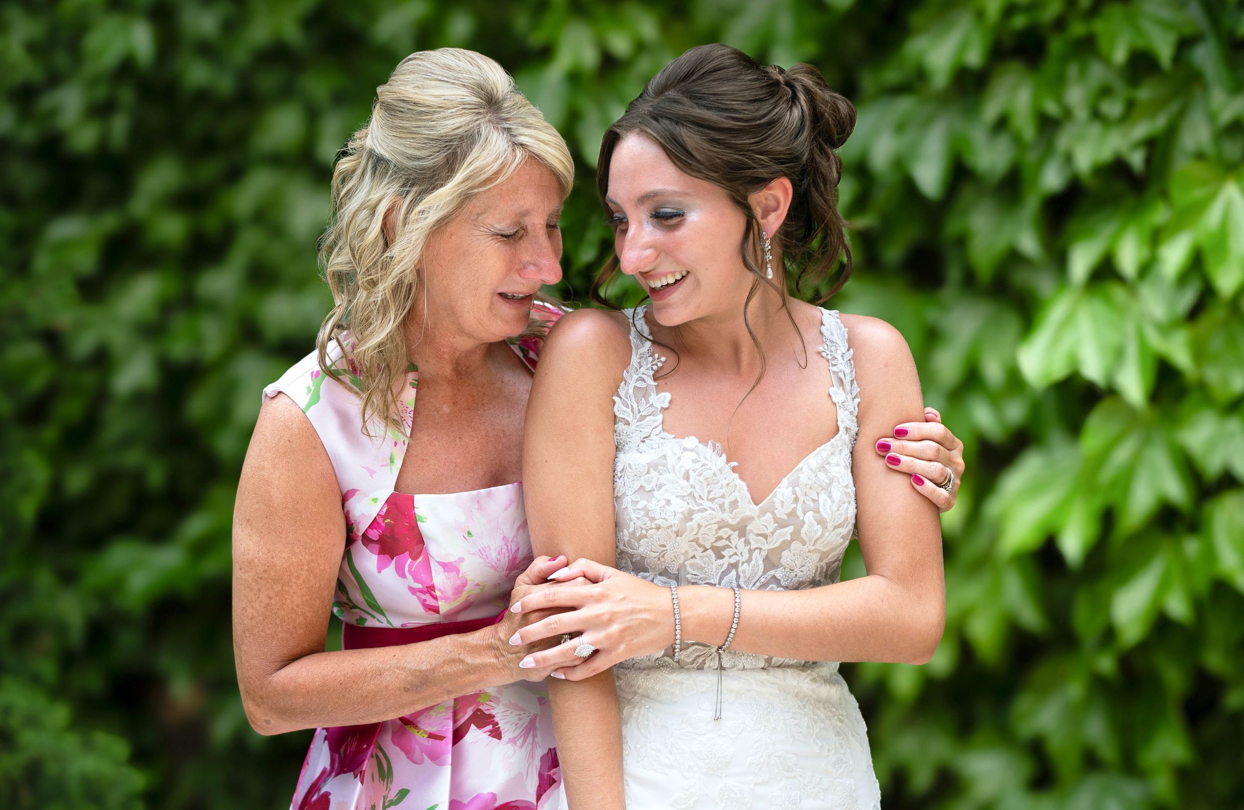 Bride in lace gown shares a tender moment with older woman in floral dress, laughing together outdoors.