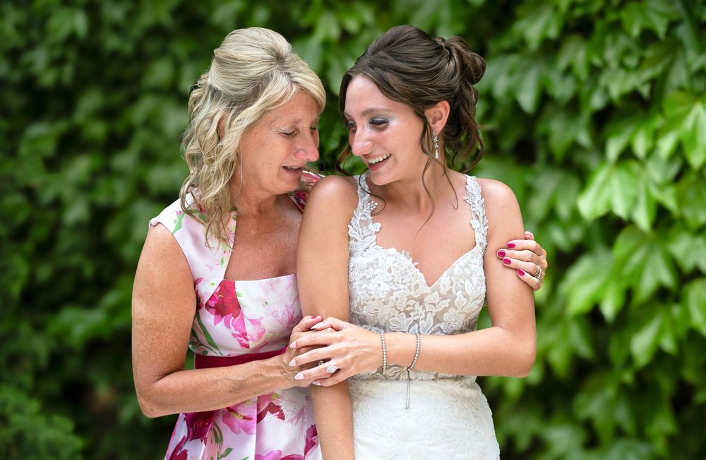 Bride in lace gown shares a tender moment with older woman in floral dress, laughing together outdoors.