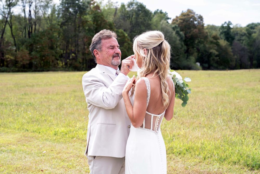 Father and bride share emotional first look moment in sunlit meadow, groom in tan suit, bride in white lace gown.