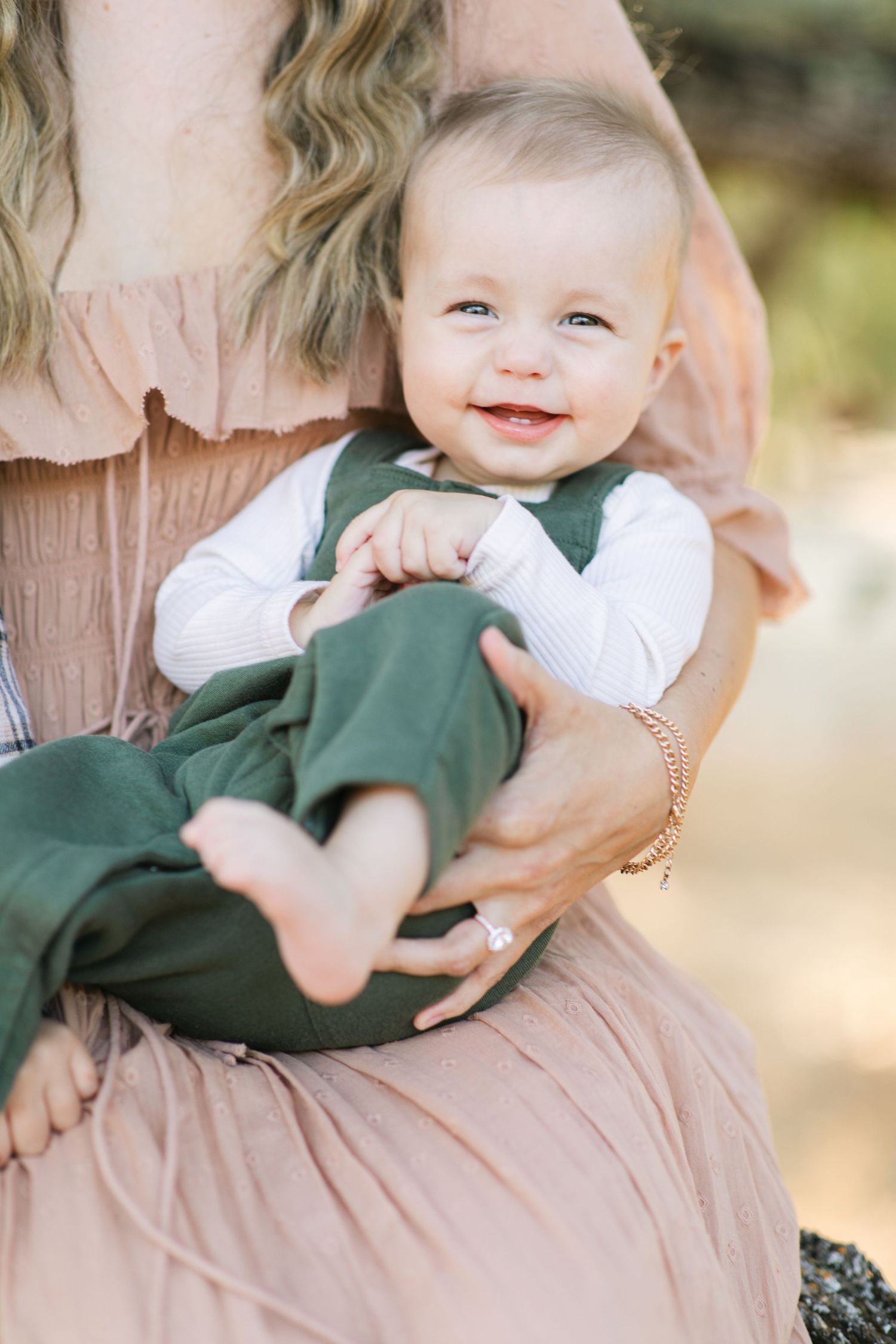 Smiling baby in green overalls and white shirt being held by a woman in a pink dress outdoors.