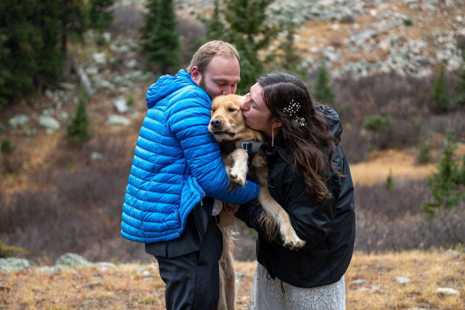 Couple in outdoor winter setting kissing a golden retriever puppy, surrounded by mountain scenery.