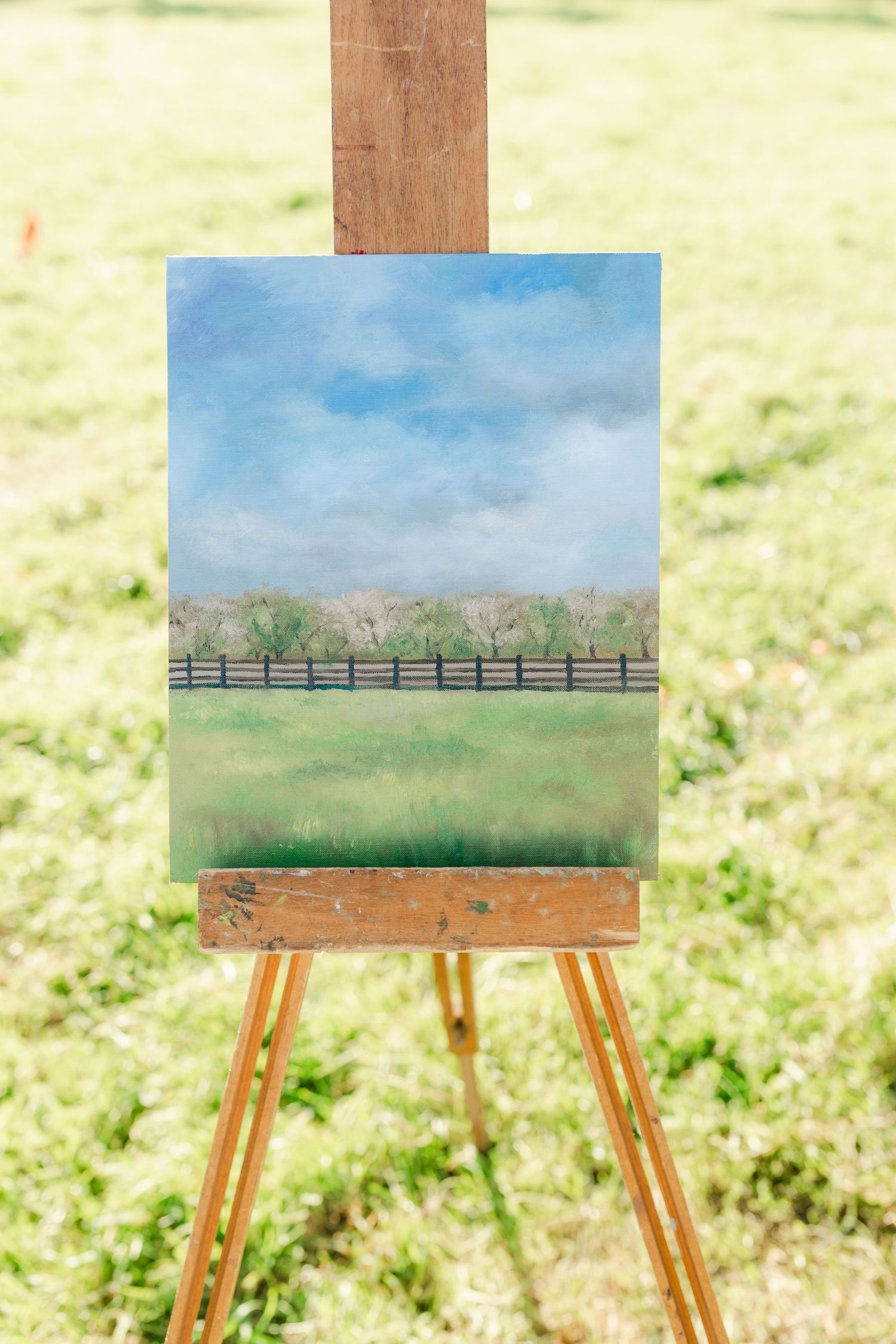 A landscape painting on a wooden easel outdoors, depicting a green field with a fence and trees under a cloudy sky.