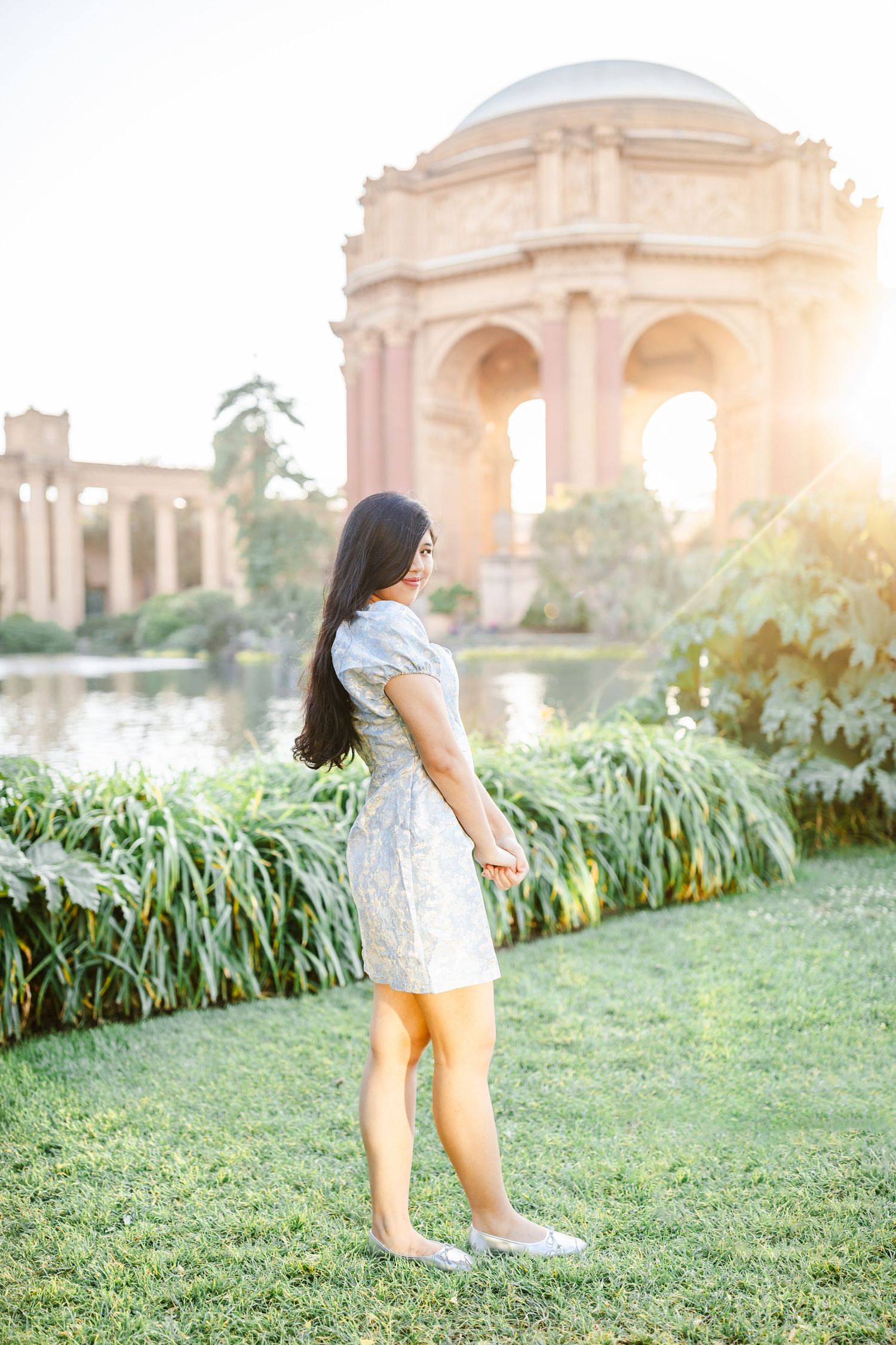 Young woman in silver dress stands on grass before the Palace of Fine Arts rotunda at golden hour in San Francisco.