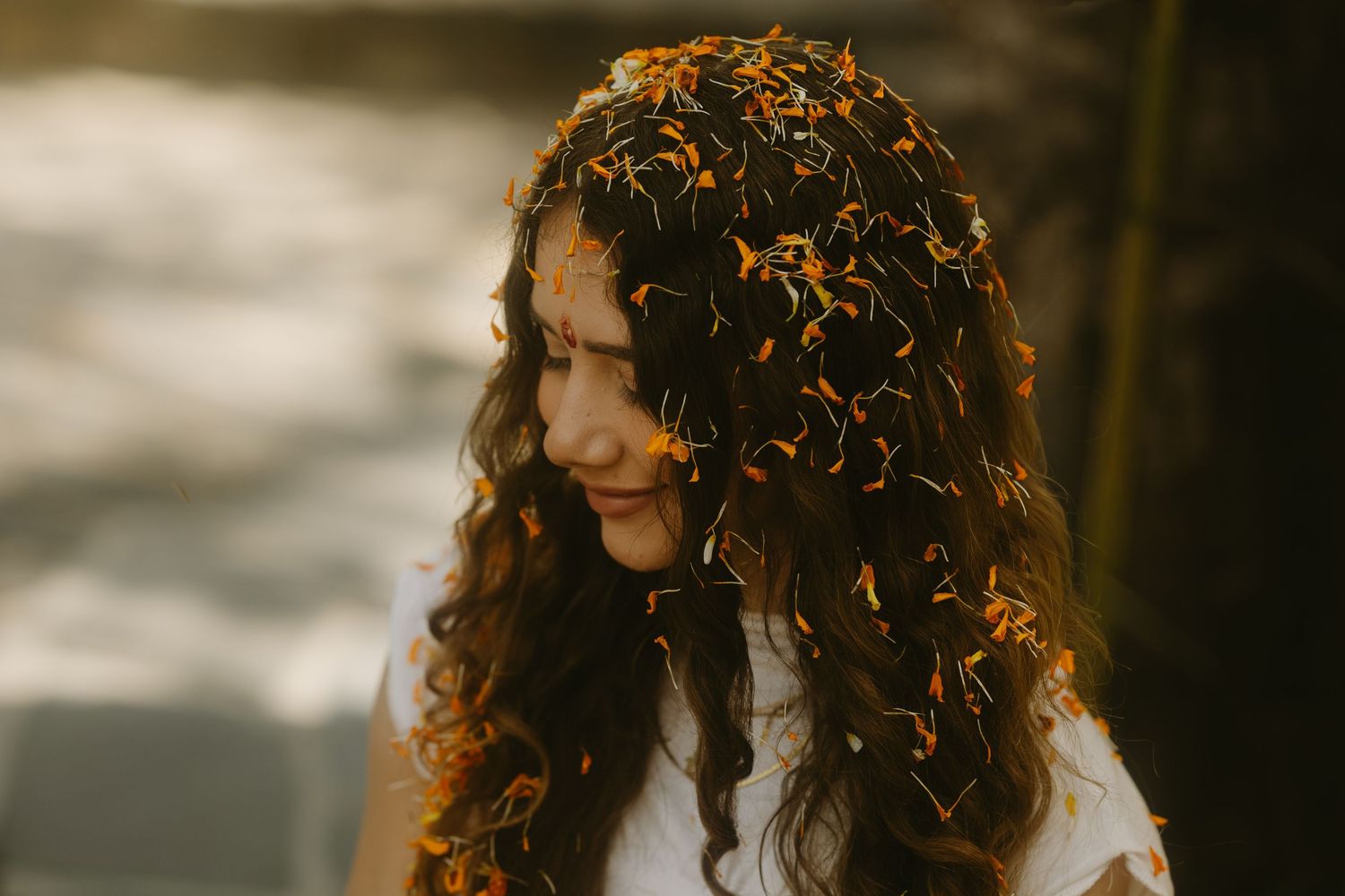 Young woman with curly hair covered in orange flower petals smiling in golden sunlight.