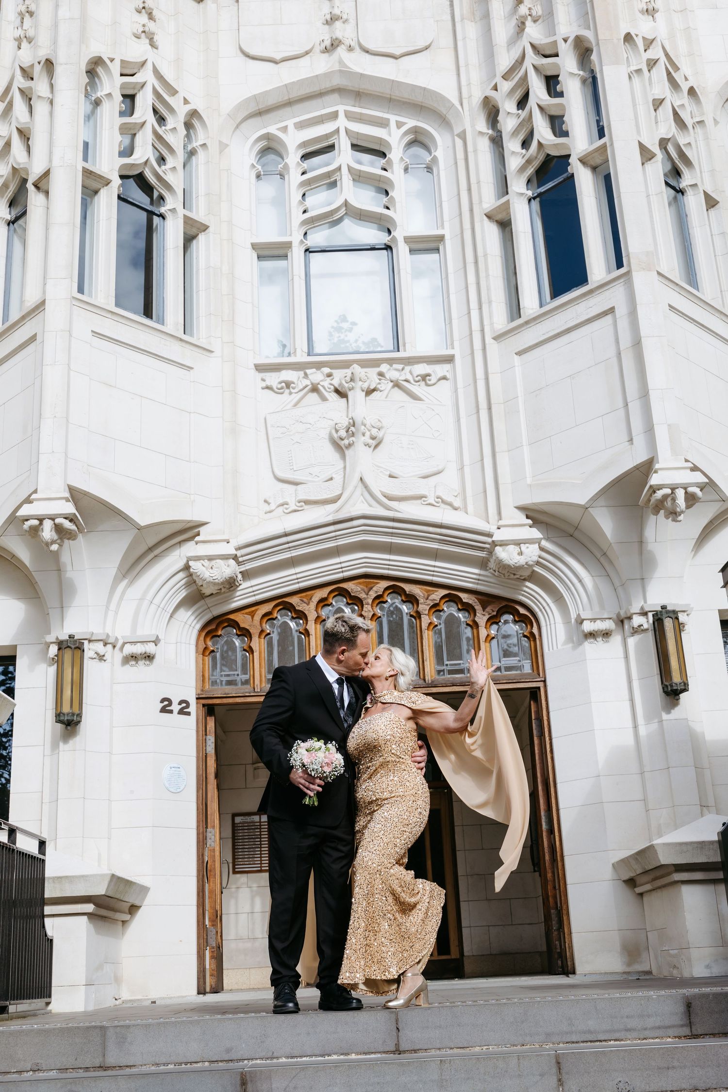 Couple kissing on steps of ornate white building, bride in gold gown holding bouquet, groom in black suit.