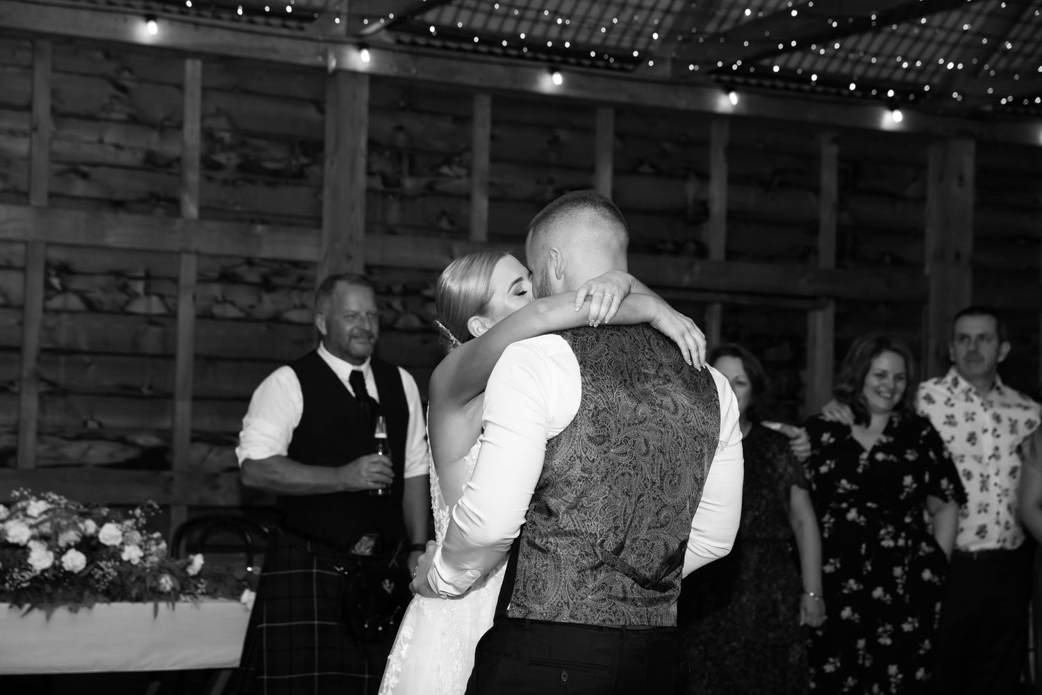 Black and white photo of a couple sharing their first dance at a rustic barn wedding reception with guests watching.