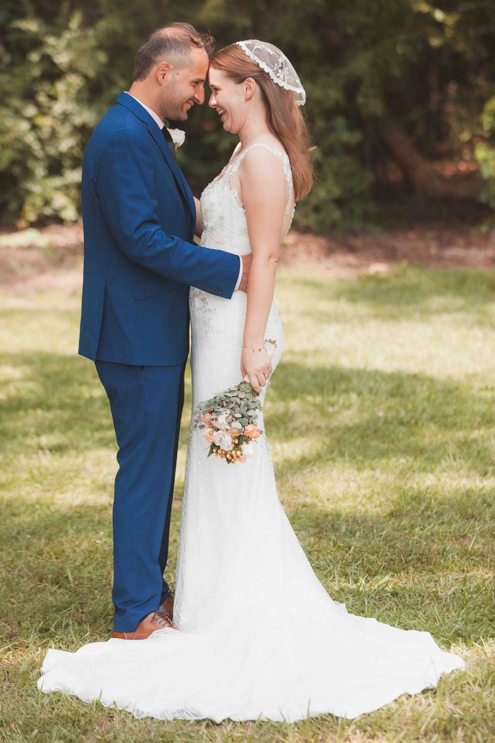 Bride and groom smiling at each other in a sunny garden, bride holding a delicate floral bouquet.