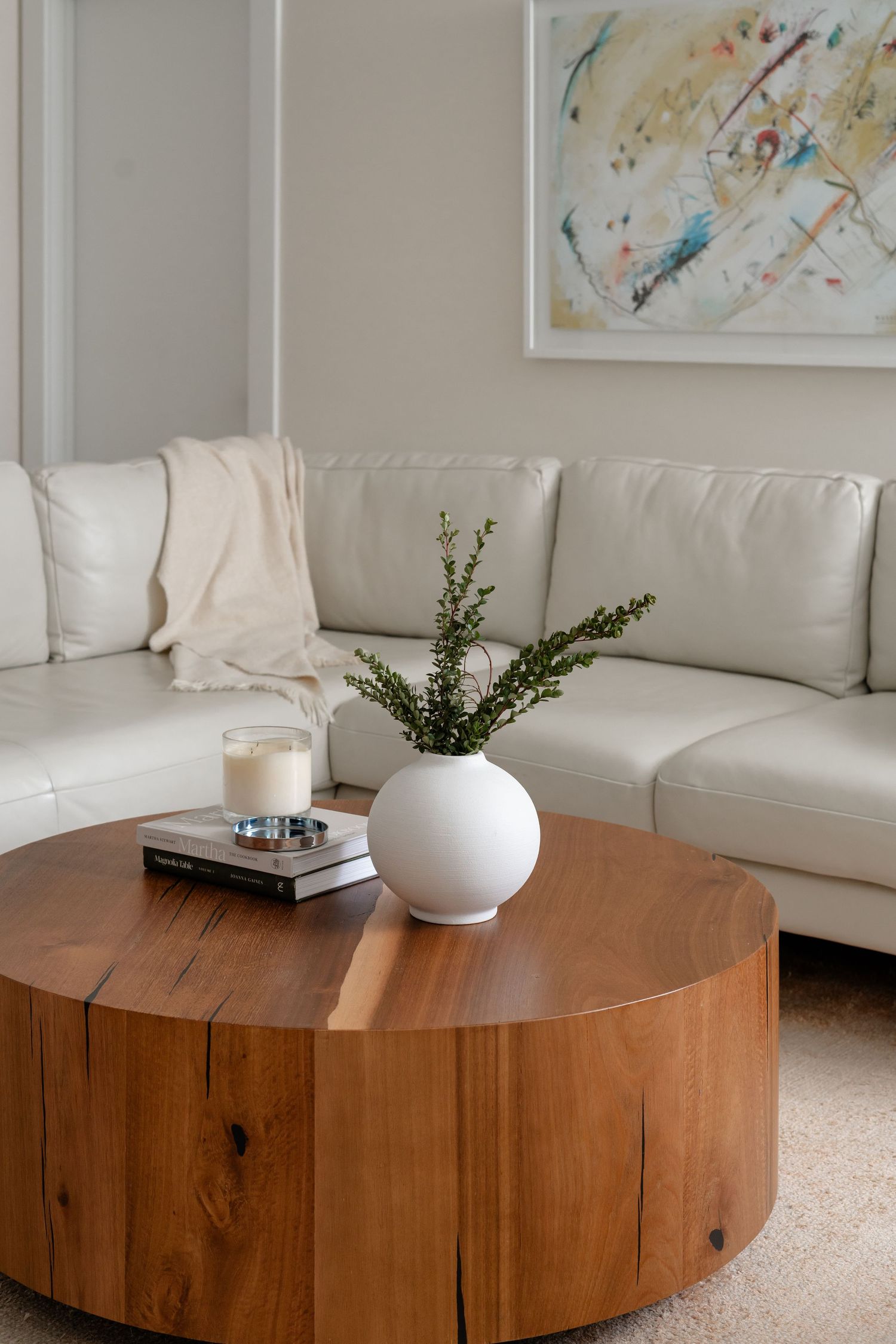 Modern living room with round wood coffee table, white vase, candle, and cream leather sectional sofa with abstract art.