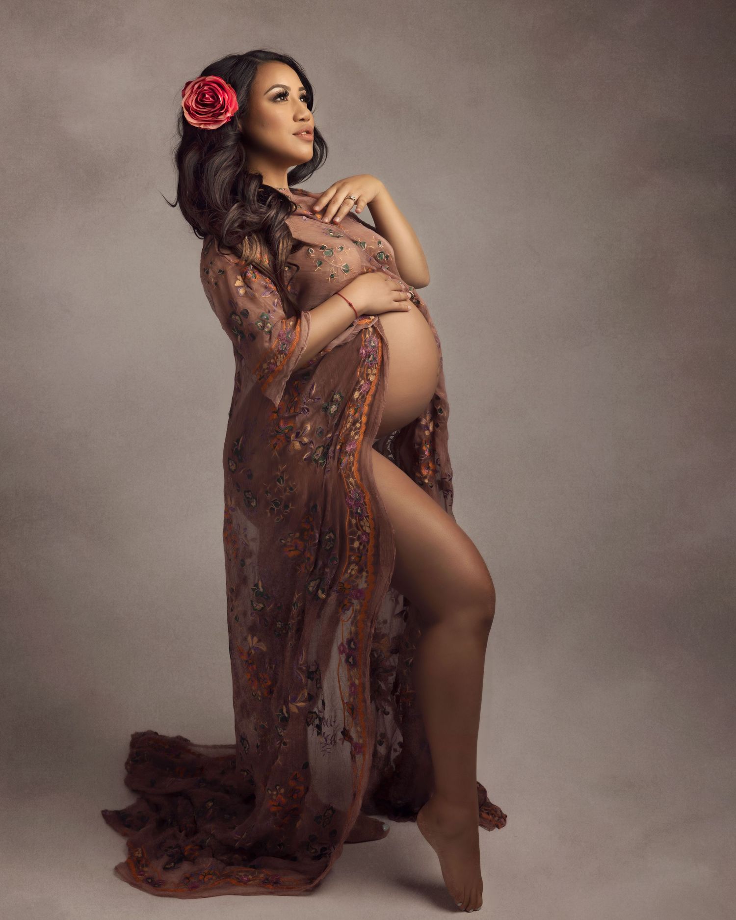 Pregnant woman in flowing brown lace maternity gown with red flower hair accessory poses gracefully in studio.