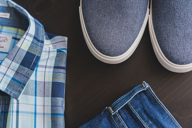 Flat lay of a blue plaid shirt, denim jeans, and gray slip-on sneakers on a dark wooden surface.