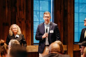 Man in blazer speaks at indoor event with microphone, woman stands behind him in rustic wood-paneled venue.
