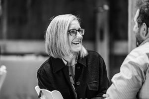 Black and white photo of a smiling woman with short blonde hair and glasses laughing at a social gathering.